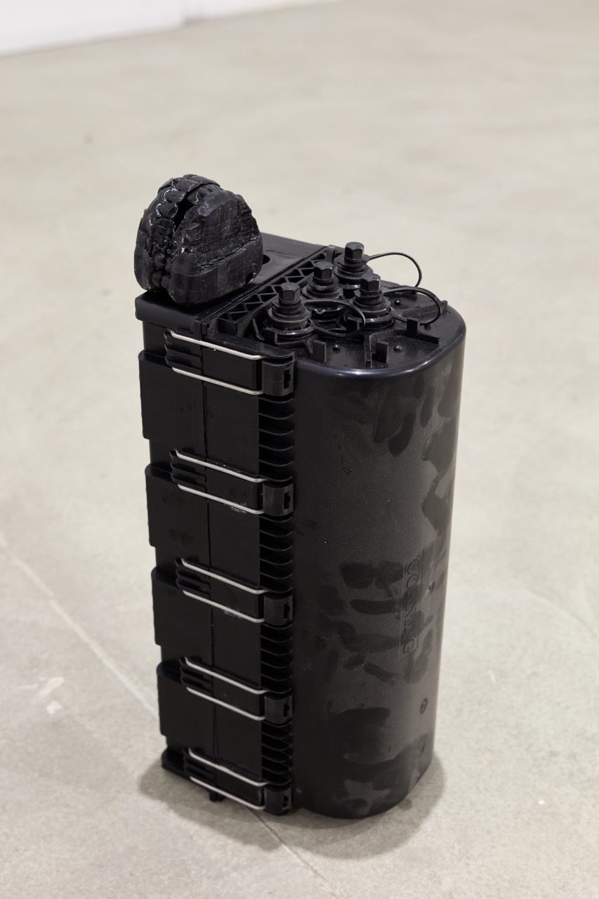 Black ribbed cylindrical object with a dark sculptural form resting on top, displayed on a pale floor.