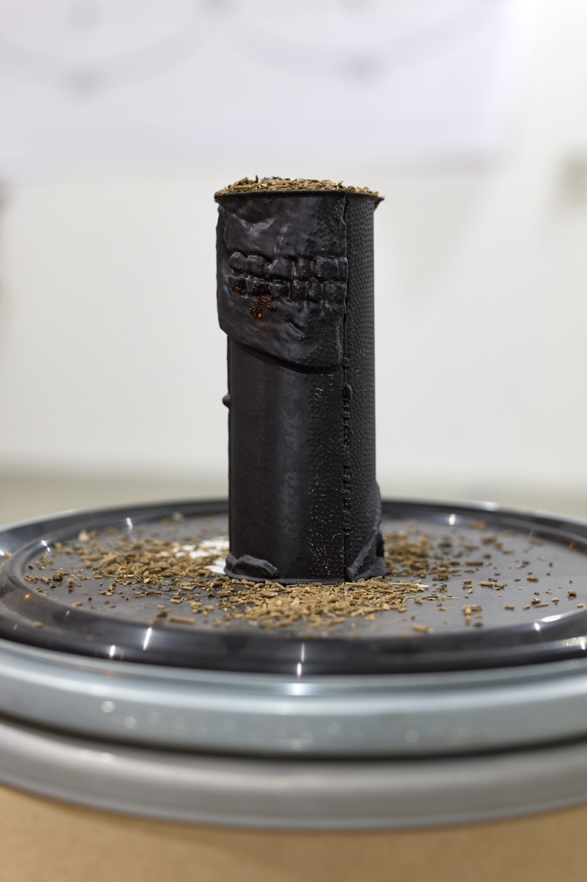 Tall black cylindrical sculpture on a metal tray scattered with dried plant material.