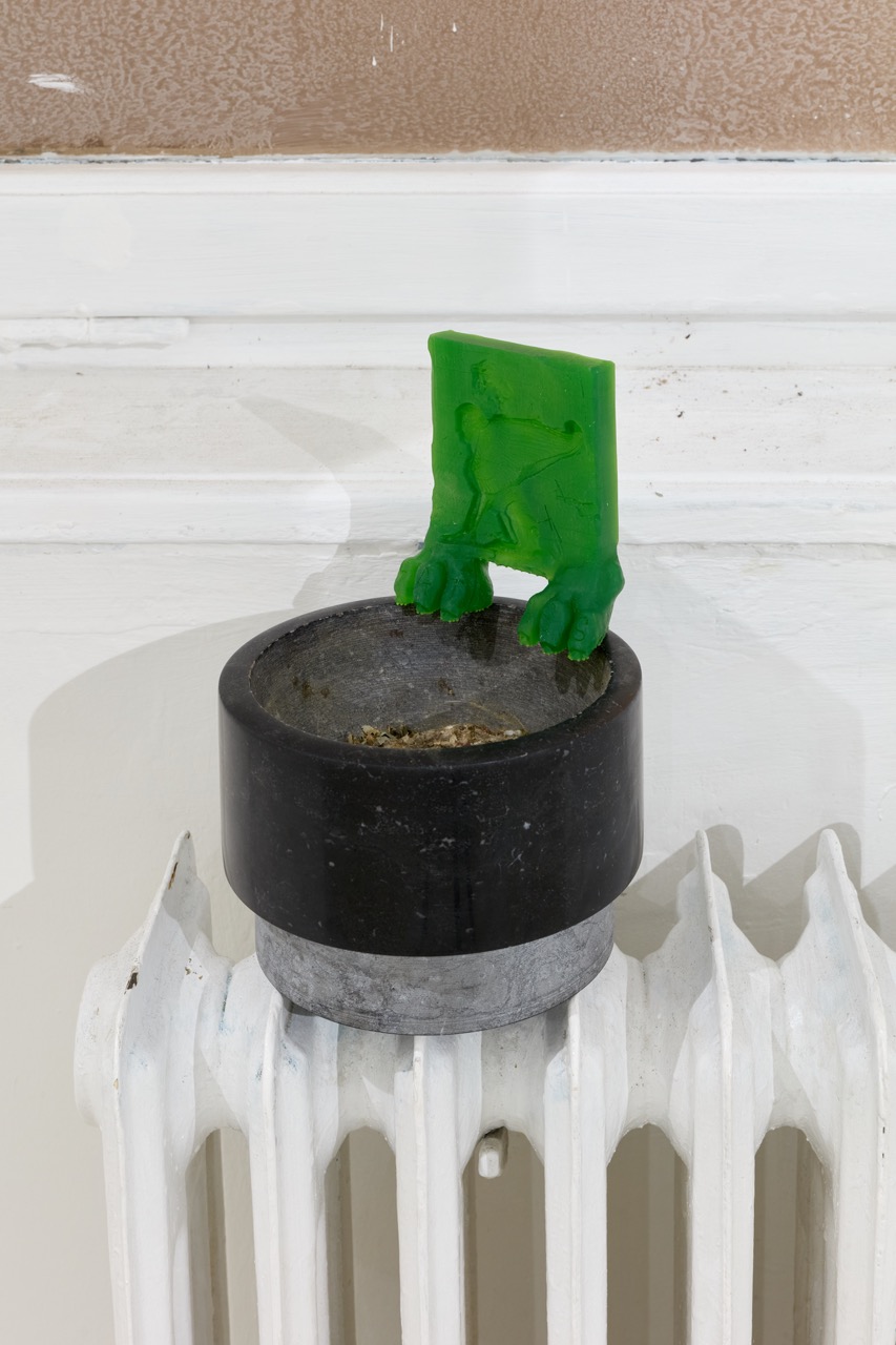 Bright green sculptural form perched on a black circular vessel above a white radiator.