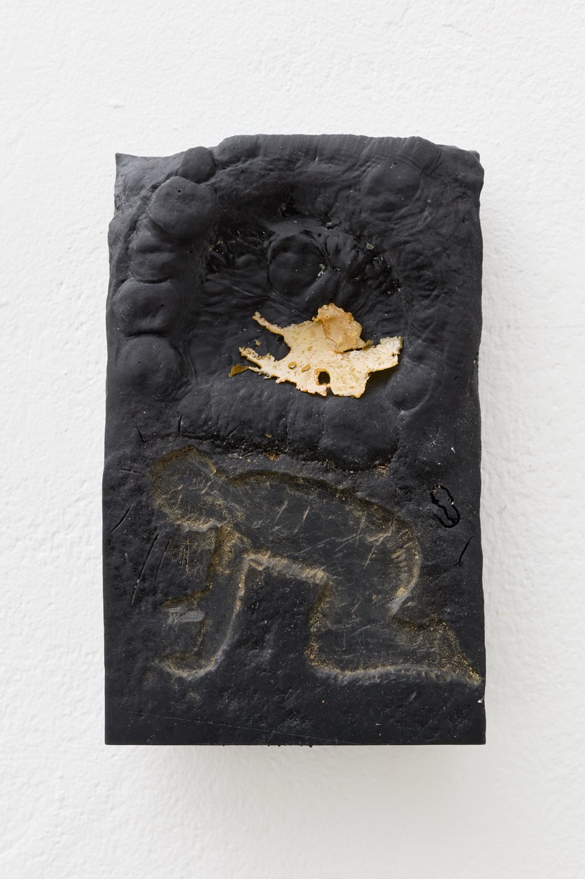 Black sculptural relief with gold and pale markings, mounted on a white wall.