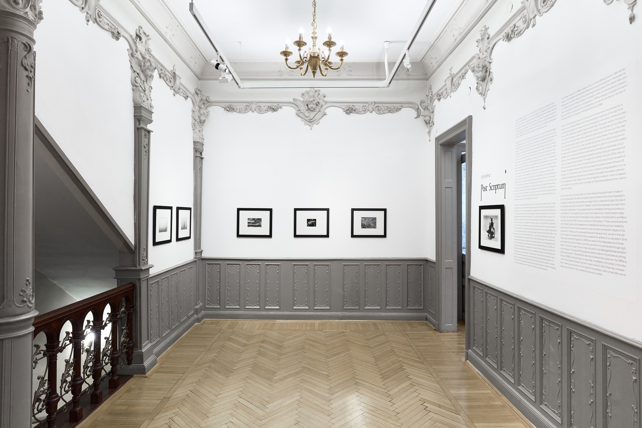 A wide view of the Post Scriptum exhibition with framed photographs, decorative grey wall panels, a chandelier and a staircase.
