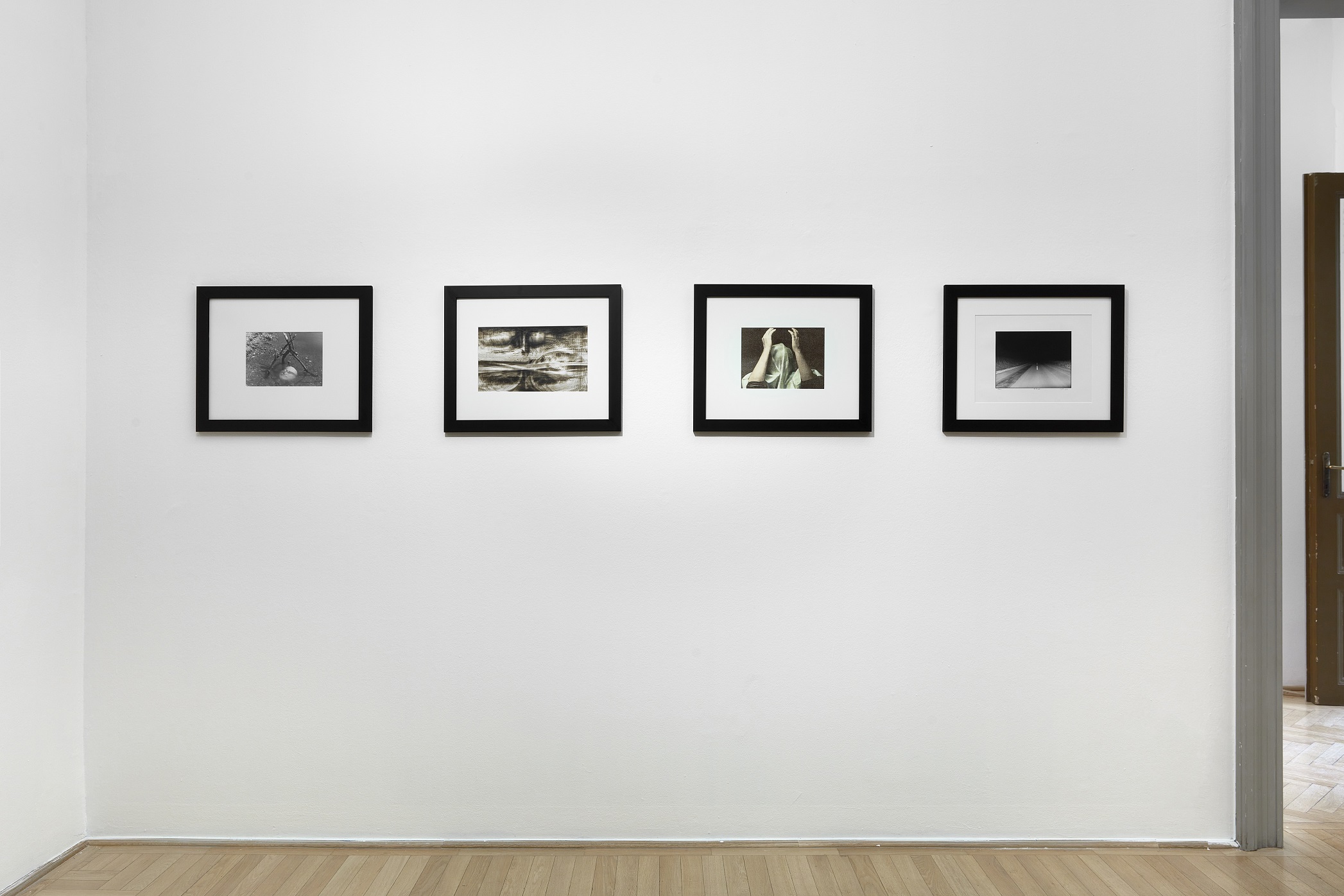 Four framed photographs from Post Scriptum hang in a row on a white gallery wall beside an open doorway and parquet floor.